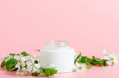 Beyaz çiçekli pembe arka planda beyaz kavanozda yüz kremi. Doğal kozmetik, organik güzellik kavramı. Boşluğu kopyala