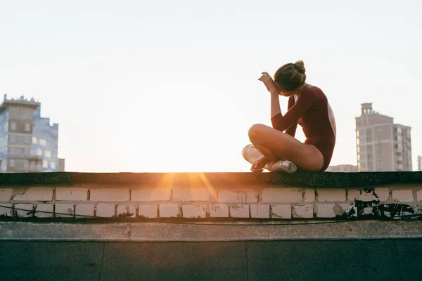 young and graceful ballerina sitting on roof of city building at sunset