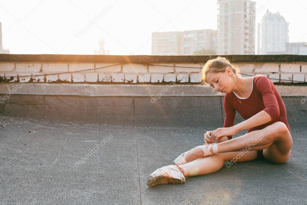 young and graceful ballerina sitting on roof of city building at sunset and tying point shoes