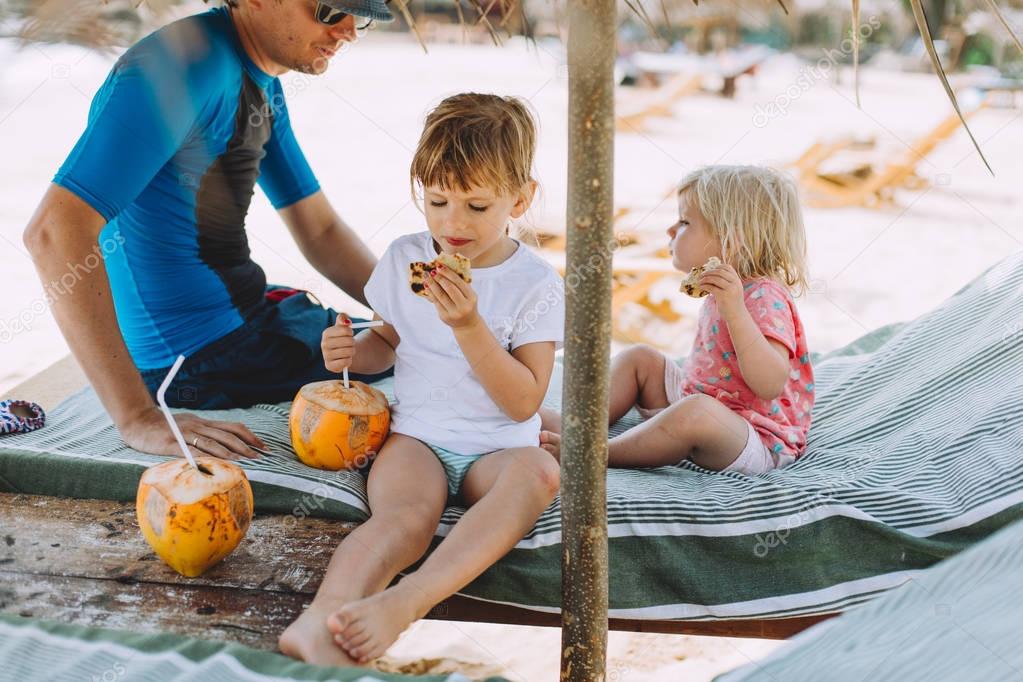 two cute little sisters drinking coconut and their father at beach 