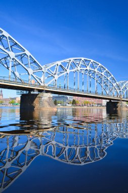 Riga şehrindeki nehir boyunca uzanan metal köprü ve onun saf mavi sudaki yansıması..