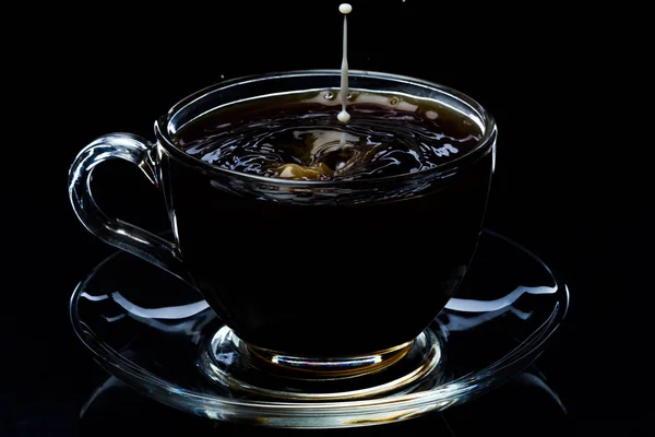 Drops of milk fall into a glass cup with black coffee, black background