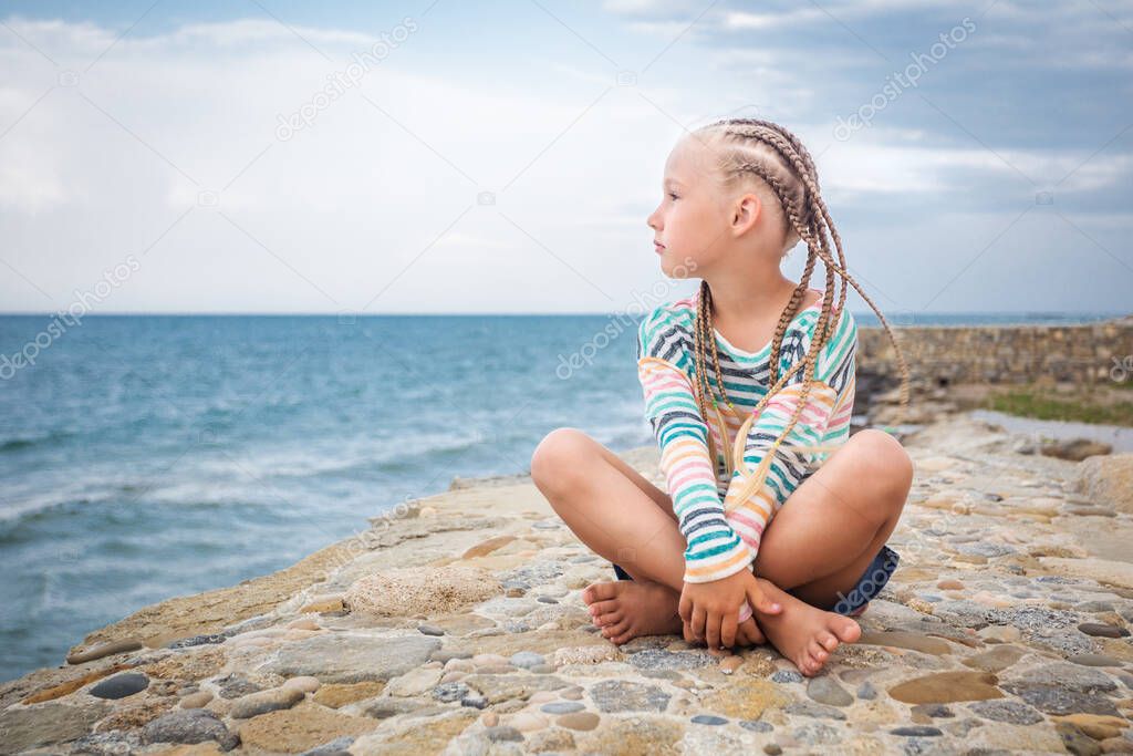 A little girl is sitting on the rocks by the sea. Long braided hair. Sea coast. Stare into the distance. Great stones. Summer evening. Dreams of good things. It's good to be alone.