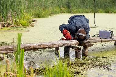 Mavi üniformalı bir kaynakçı, kaynak maskesi ve koruyucu kırmızı eldivenler, kaynak makinesi ile metal kaynaklama, bataklıktaki tahta köprü..