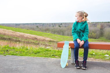 Yeşil ceketli bir kız bankta oturuyor. Tıbbi maskeli bir kız. Mavili kaykay. Arka planda orman var. Covid-19 Coronavirüs. Evde kal..