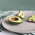 On a plate is a ripe avocado and a sandwich of cereal bread, soft cheese and slices of avocado. Tablecloth with palm leaves, green wall. Proper nutrition, vegetarian. Breakfast for coffee. Light snack