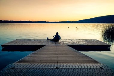 Günbatımı ve Gölü izlerken bir ahşap platform üzerine oturan adam 