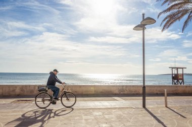 Arka planda Akdeniz varken güneşli bir sabahta Cala Millor 'ın gezinti güvertesinde çalışan bisikletçi. Mallorca Adası