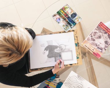 Sanatçı cam masanın üzerinde yer alan resimli çocuk kitabının ikinci baskısı için kalem çizimi yapıyor. Yaratıcı ortam