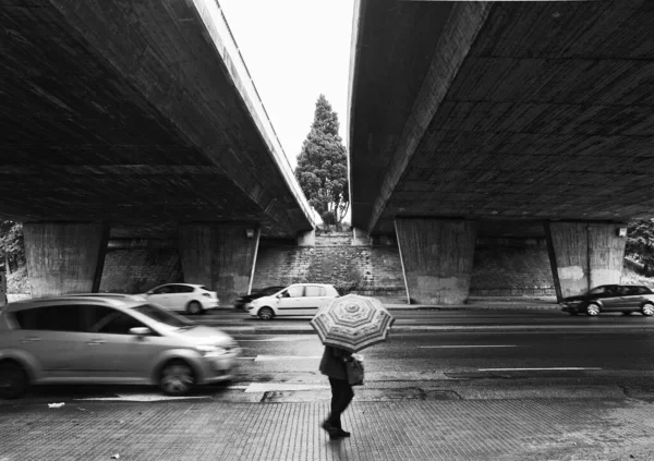 Person walking with umbrella under two concrete bridges and in the background the moving car traffic. Urban image in black and white