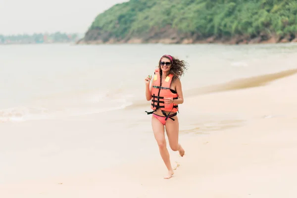 Glückliches Mädchen läuft in orangefarbener Schwimmweste am Strand in der Nähe der Brandung entlang — Stockfoto