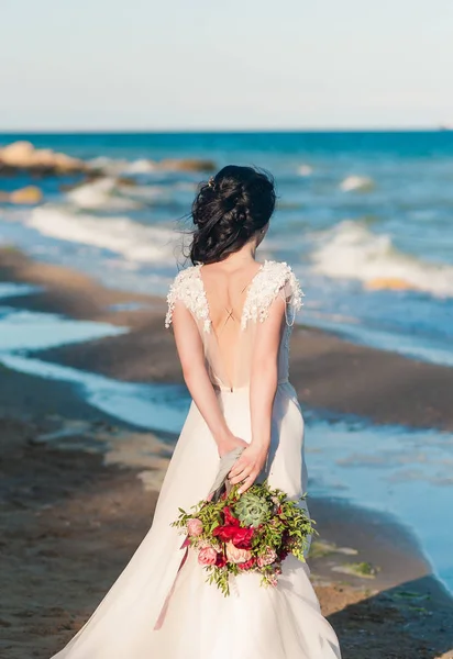 Beautiful bride in luxury wedding dress with bouquet at the sea side. Wedding by the sea. Bride walking around the sea near the place of the wedding ceremony. Back view