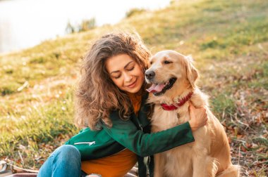 Gülümseyen bir kadın köpeğine sarılıyor. Golden Retriever köpeği, güneşli bir günde dışarıda yürüyen kıvırcık bir kadınla oynuyor. Sevgi ve evcil hayvan için bakım.