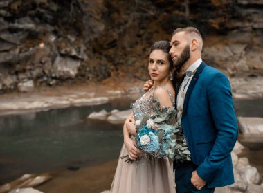 Dağ nehrinin yanındaki düğün gününde güzel bir çift gelin ve damat. Mutlu evlilik çifti taşların üzerinde oturuyor, yumuşak güneşli ışıklar. Evlilik gününde sevecen erkek ve kadın.