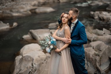 Dağ nehrinin yanındaki düğün gününde güzel bir çift gelin ve damat. Mutlu evlilik çifti taşların üzerinde birbirini kucaklıyor, yumuşak güneşli ışıklar. Karpat Dağlarında Düğün.