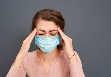 Tıbbi maskeli genç esmer kız kafasını gri arka planda tutuyor. Kadın şiddetli baş ağrısı ve migren yüzünden acı çekiyor. Duygularla yüzleş.