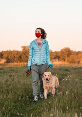 Yüzünde tıbbi maske ve ellerinde koruma eldiveni olan Golden Retriever köpeğiyle temiz havada yürüyen bir kız. Koronavirüs salgını, karantina.