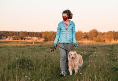 Yüzünde kırmızı koruyucu maske ve ellerinde tıbbi eldivenler olan Golden Retriever köpeğiyle temiz havada yürüyen bir kız. Koronavirüs salgını, karantina.