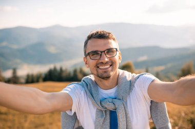 Yakışıklı sakallı adam selfie çekiyor. Gezgin Adam bir dağ manzarasının kamera arkasında gülümser. Yolculuk Konsepti