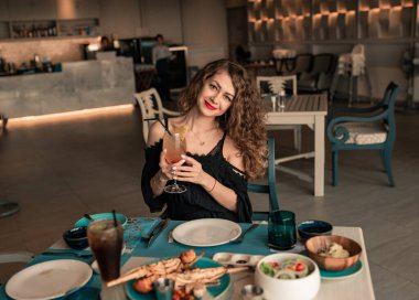 Portreyi kapat. Kıvırcık saçlı seksi esmer kadın lüks yaz barında eğleniyor. Siyahi seksi elbiseli, renkli kokteylli çok çekici bir kız. Restoranda yemek yiyen bir kadın.