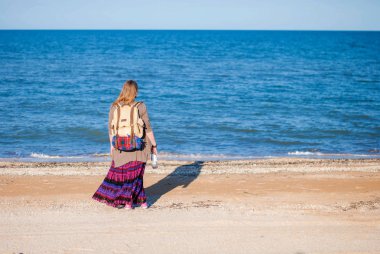 Bir kız geri duruyor ve denizde görünüyor. Hippi kız denizde görünüyor. Bir şişe şarap ile deniz kenarında bir kadındır. eski bir sırt çantası ve bir şişe şarap ile hippi kız.