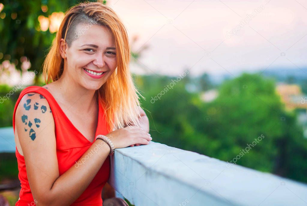 Beautiful red-haired girl in a red dress and tattooed laughs on the veranda of a summer restaurant