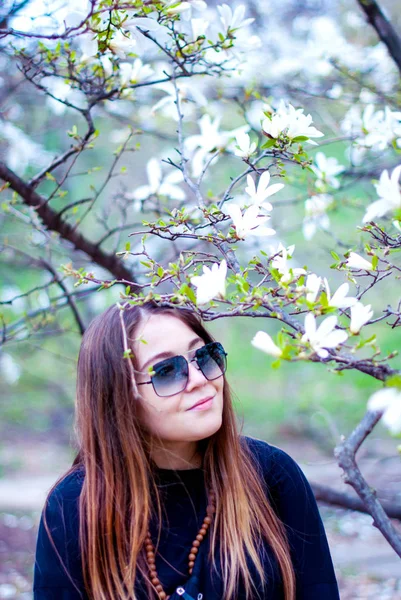 Beautiful girl in sunglasses smiling near blooming trees. Fat girl smiles in the magnolia garden. Girl with long hair wearing sunglasses.