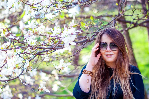Beautiful girl in sunglasses smiling near blooming trees. Fat girl smiles in the magnolia garden. Girl with long hair wearing sunglasses.