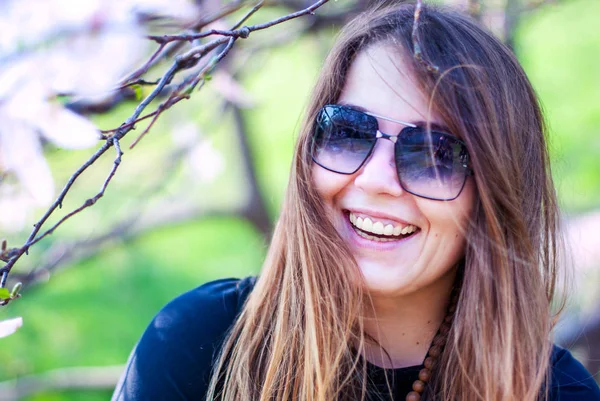 Vacker flicka i solglasögon leende nära blommande träd. Fett flicka leenden i magnolia trädgården. Tjej med långt hår bär solglasögon. — Stockfoto