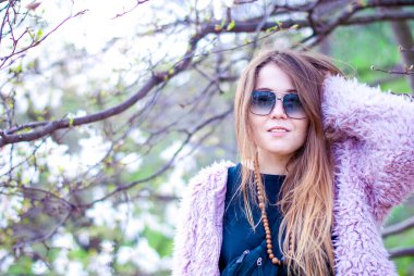 Güzel kız yapay bir pembe ceket. Kız uzun saçlı güneş gözlüğü takıyor. Kadın gülümsüyor güneş gözlüğü ve Manolya bahçedeki pembe ceket.