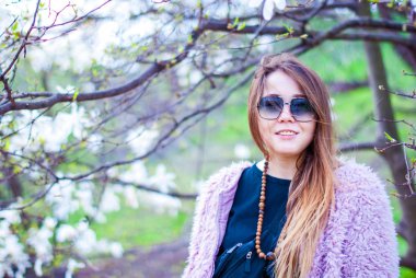 Güzel kız yapay bir pembe ceket. Kız uzun saçlı güneş gözlüğü takıyor. Kadın gülümsüyor güneş gözlüğü ve Manolya bahçedeki pembe ceket.