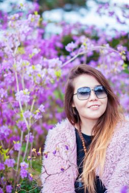 Kız bir yapay kürk manto. Güneş gözlüklü kız gülümsüyor. Mutlu kadın çiçek açması ağaçlar yakınındaki pembe bir ceket.