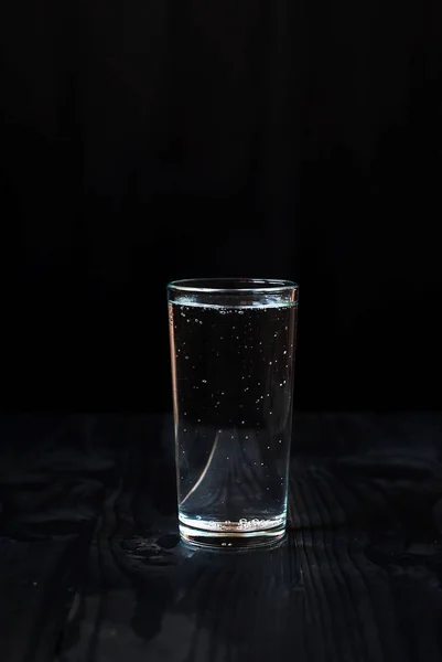 Hanya segelas air dengan latar belakang hitam. Air mineral dalam gelas di atas meja kayu gelap . — Stok Foto