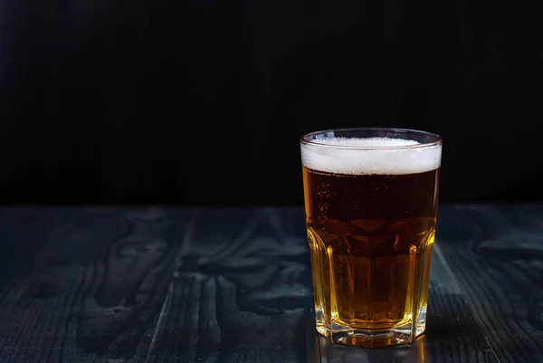 Just a glass of beer on a dark background. A glass of light beer on a wooden table.