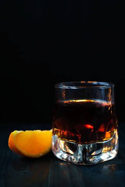 Bir kadeh viski ya da siyah bir arka plan ve ahşap bir masa üzerinde turuncu bir dilim ile konyak