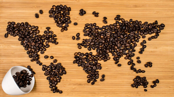 Map of the world made of roasted arabica coffee beans laying on bamboo wooden textured background with white ceramic cup full of scattered roasted coffee beans