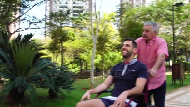 Vader wandelend met gehandicapte zoon in rolstoel op park — Stockvideo