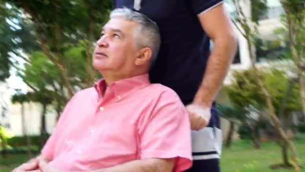 Fils marchant avec un père handicapé en fauteuil roulant au parc — Video