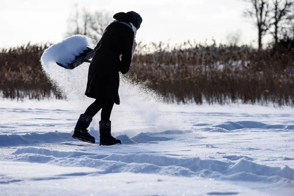 Giovane donna in cappotto nero pulizia ghiaccio dalla neve. lancio di neve — Foto Stock