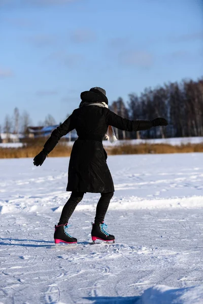 Giovane donna in cappotto nero pattinaggio sul lago ghiacciato nella neve, le — Foto Stock