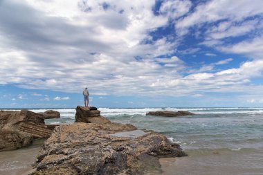 Deniz kıyısı genç adam maliyetlerinde. Kot şort ve beyaz keten çanta bir omuz üzerinde yalınayak genç adam bulutlu bir uçan hava kumlu plajında maliyeti ve Uzaktan görünüyor