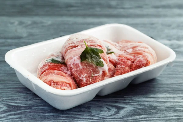 Frozen meat in hoarfrost in a white plastic tray with greens.