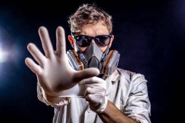 Koyu güneş gözlüklü beyaz erkek, yarım yüz solunum cihazı ya da gaz maskesi takıyor, kameraya doğru uzanıyor, karanlık arka planda tozlu ortamda dramatik ışıklandırma efektleri var. Virüs konsept korkusu.