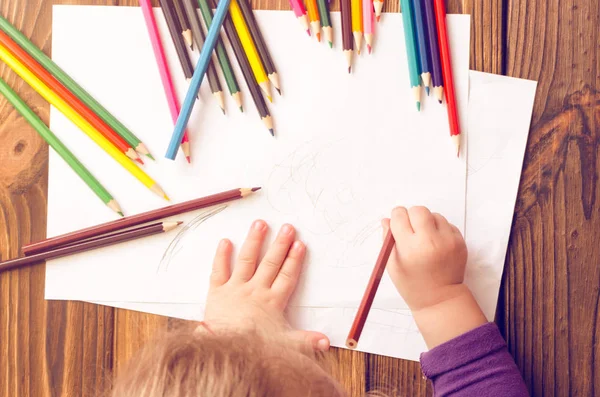 Çocuğun elleri ahşap bir masa üzerinde beyaz kağıda renkli kalemler ile boyanır