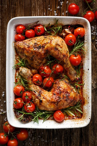 Baked chicken quarter with cherry tomatoes and rosemary in a casserole dish on wooden table