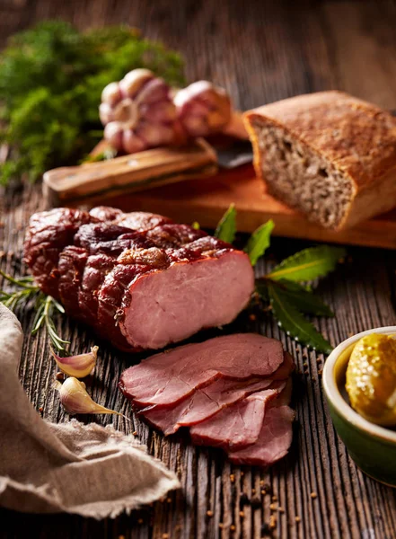 Smoked ham sliced on a wooden rustic table with addition of fresh aromatic herbs and spices.  Natural product from organic farm, produced by traditional methods