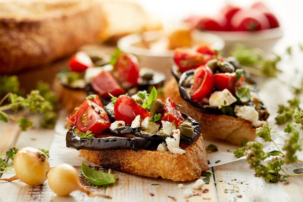 Bruschetta com beringela grelhada, tomate cereja, queijo feta, alcaparras e ervas aromáticas frescas, sobre uma mesa de madeira. Delicioso aperitivo mediterrânico — Fotografia de Stock