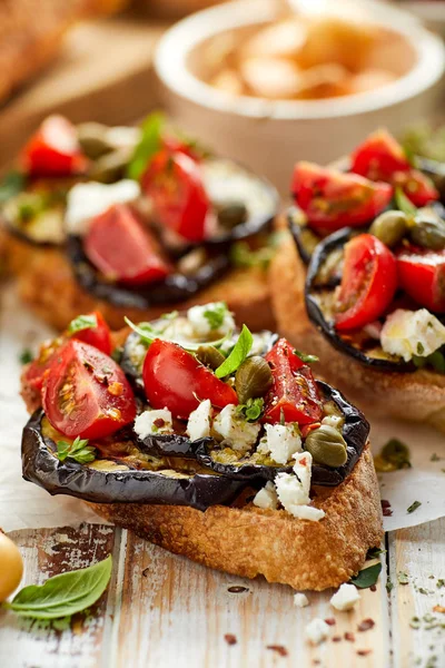 Bruschetta con berenjena a la parrilla, tomates cherry, queso feta, alcaparras y hierbas aromáticas frescas, sobre una mesa de madera. Delicioso aperitivo mediterráneo — Foto de Stock