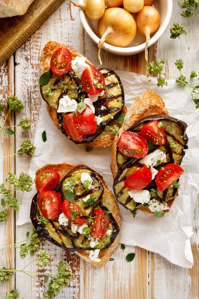 Bruschetta with grilled eggplant, cherry tomatoes, feta cheese, capers and fresh aromatic herbs on a wooden table. Delicious Mediterranean appetizer — Stock Photo, Image