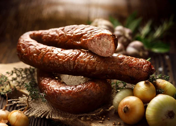 Wędzona kiełbasa na drewnianym rustykalnym stole z dodatkiem świeżych aromatycznych ziół i przypraw, produkt naturalny z gospodarstwa ekologicznego, produkowany tradycyjnymi metodami — Zdjęcie stockowe